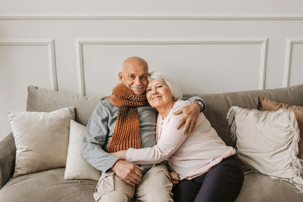 Joyful elderly couple sitting on a sofa, smiling and hugging, expressing their happiness with the domiciliary care services provided by SWL Care Haven caregivers.
